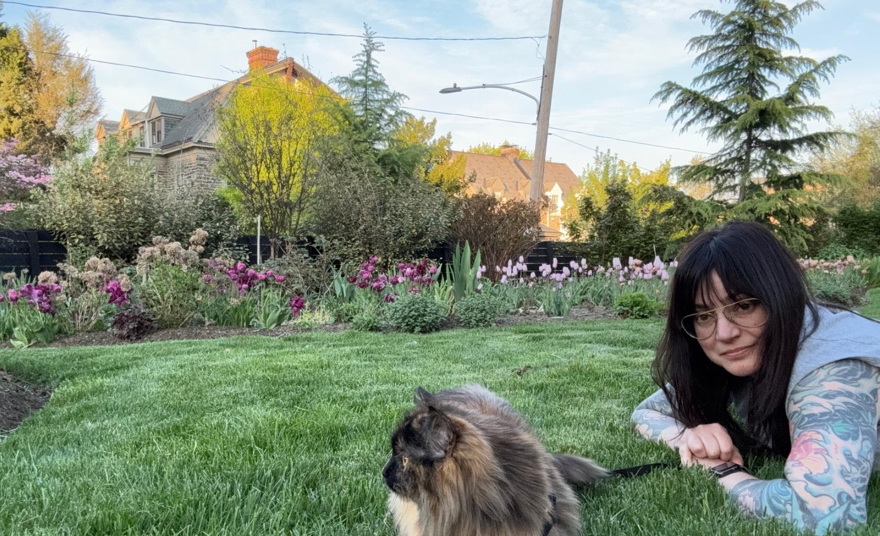 Sullivan Owen in her home garden in Philadelphia, lying on the grass next to her long-haired cat Chewie during peak spring bloom season.